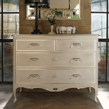 The Classic Louis Style Chest Of Drawers, featuring an elegant off-white finish and ornate handles, stands against a wall with large windows. Decoratively placed on top are a black lamp with a patterned shade, a small decorative mirror, and two white ceramic elephant figurines.