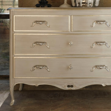 The Classic Louis Style Chest of Drawers, with its ornate handles and gold accents, stands against a light background. It features three large drawers at the bottom and two smaller drawers at the top, all adorned with decorative handles. This vintage wooden dresser is elegantly positioned on a rustic wooden floor.