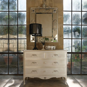 A Classic Louis Style Chest Of Drawers with ornate handles sits against a large window. Above it is a decorative mirror with intricate details. On the dresser, there is a black lampshade and a small elephant figurine. The background reveals an outdoor garden through the window.