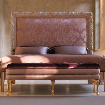 Large Classic Louis XVI Style Bench A luxurious bed featuring an ornate pink and gold headboard, along with a matching bedspread and pillows, is complemented by the Large Classic Louis XVI Style Bench positioned at the foot. The bench boasts a tufted velvet cushion on top and elegant gold claw-foot legs. This setting is situated in a warmly lit, sophisticated room.
