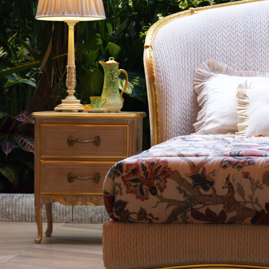 A luxurious bedroom scene featuring the **Classic Ornate Two Drawer Bedside Cabinet**, adorned with a decorative lamp and a green ceramic teapot. A section of a plush, upholstered bed with intricate floral bedding and a ruffled pillow rests adjacent to the cabinet. Lush plants are in the background.