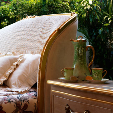 Classic Ornate Two Drawer Bedside Cabinet Transform a cozy corner with an elegant upholstered chair featuring a white and gold woven backrest and a floral cushion, positioned beside a wooden table. The table is accompanied by the Classic Ornate Two Drawer Bedside Cabinet, which holds a green, hand-painted china tea set. This charming scene is further enhanced by lush greenery in the background.