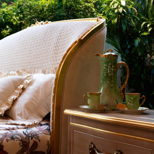 Transform a cozy corner with an elegant upholstered chair featuring a white and gold woven backrest and a floral cushion, positioned beside a wooden table. The table is accompanied by the Classic Ornate Two Drawer Bedside Cabinet, which holds a green, hand-painted china tea set. This charming scene is further enhanced by lush greenery in the background.