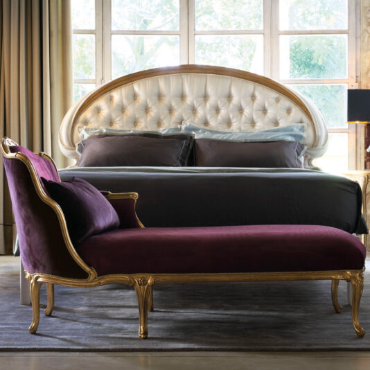 A luxurious bedroom features a Classic Style Button Upholstered Bed adorned with dark bedding, positioned against expansive windows. In the foreground, a purple velvet chaise lounge with gold detailing sits on a gray rug, adding an elegant touch to the space.