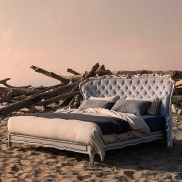 Classic Style Button Upholstered Winged Bed A Classic Style Button Upholstered Winged Bed, complete with plush pillows and a cozy comforter, is elegantly arranged on a sandy beach where driftwood is scattered in the background. The scene is bathed in a soft peach hue from the sky, hinting at either sunrise or sunset.