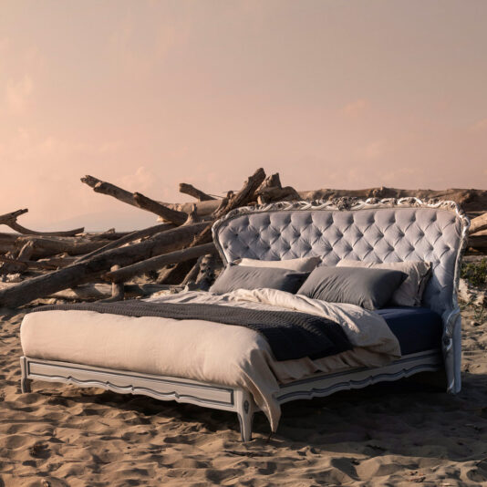 A Classic Style Button Upholstered Winged Bed, complete with plush pillows and a cozy comforter, is elegantly arranged on a sandy beach where driftwood is scattered in the background. The scene is bathed in a soft peach hue from the sky, hinting at either sunrise or sunset.