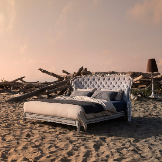 The Classic Style Button Upholstered Winged Bed, featuring a luxurious white tufted headboard, is positioned on a sandy beach amidst driftwood under a pinkish sky. The bed is neatly made with white and dark-toned bedding, while a tall, dark standing lamp blends into the serene beachfront setting.