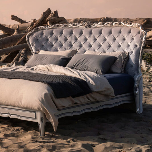 A Classic Style Button Upholstered Winged Bed with a high, tufted headboard and elaborate carvings sits on a sandy beach. The bed is adorned with beige and gray bedding, with a folded dark blue blanket at the foot. Driftwood lies scattered in the background under a cloudy sky.