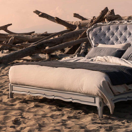 The Classic Style Button Upholstered Winged Bed is adorned with lavish linens and set on a sandy beach, surrounded by large driftwood pieces under soft, warm light.