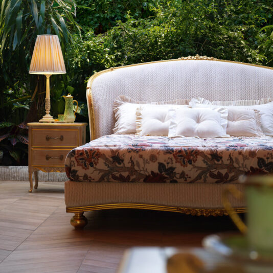 A luxurious outdoor patio features a plush white sofa with tufted cushions, a floral-patterned throw, and a golden frame. Beside it is an elegant wooden nightstand with a classic lamp reminiscent of the detailing on the Classic Style Antique Gold Winged Bed. The background is lush with green plants, adding to the serene ambiance.