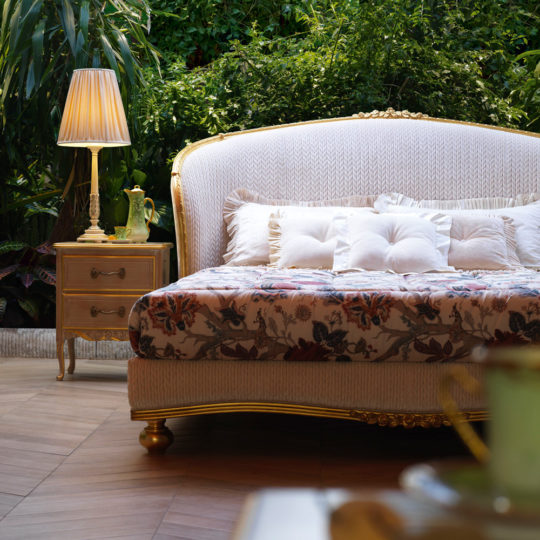 A luxurious outdoor patio features a plush white sofa with tufted cushions, a floral-patterned throw, and a golden frame. Beside it is an elegant wooden nightstand with a classic lamp reminiscent of the detailing on the Classic Style Antique Gold Winged Bed. The background is lush with green plants, adding to the serene ambiance.