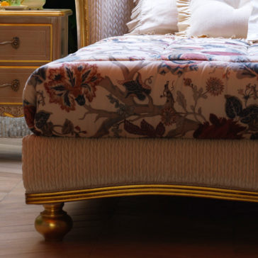 A close-up of the Classic Style Antique Gold Winged Bed highlights its elegant floral-patterned comforter and plush pillows. The intricately detailed, gold-winged bed frame adds a touch of opulence. On the left side, a wooden nightstand with a drawer can be partially seen, resting on a wooden floor.