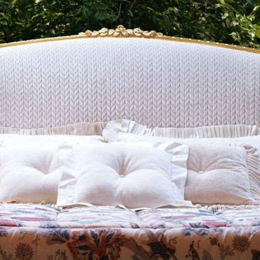 A luxurious white upholstered headboard is highlighted against a backdrop of lush green foliage, epitomizing classic style. The Classic Style Antique Gold Winged Bed is adorned with plush white pillows featuring buttoned centers and a ruffled white sheet. The quilt, patterned with colorful floral designs, perfectly complements the elegant gold-winged bed frame.