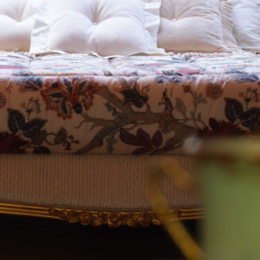A close-up view of the Classic Style Antique Gold Winged Bed adorned with a plush floral-patterned comforter and multiple white, tufted pillows. The ornate gold winged bedframe features intricate carvings and a golden finish. In the foreground, a blurred object partially obscures the lower right corner of the image.