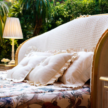 The Classic Style Antique Gold Winged Bed offers a cozy experience with its wicker headboard adorned by two white, ruffled pillows and a floral bedspread. Complementing the bed is a brass lamp with a pleated white shade on the bedside table, all set against a lush green garden backdrop.