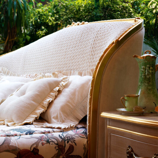 A cozy, ornately decorated bedroom features the Classic Style Antique Gold Winged Bed adorned with plush, ruffled pillows and a floral bedspread. A wooden nightstand holds a vintage tea set, framed by lush, green plants in the background, creating a serene and inviting atmosphere.