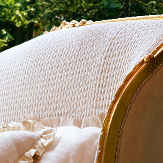 Close-up image of the backrest of an elegant, cream-colored upholstered chair with a detailed, textured fabric and gold accents. The background features lush green foliage, perfectly contrasting with the chair's classic style and ornate design reminiscent of the Classic Style Antique Gold Winged Bed.