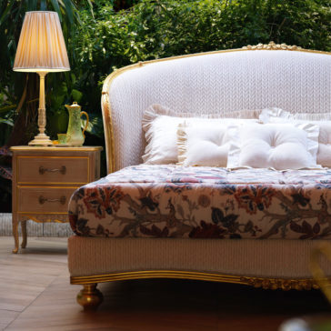A luxurious bedroom features a Classic Style Antique Gold Winged Bed with an ornate, gold-trimmed headboard and a floral-patterned bedspread adorned with multiple pillows. To the left of the bed is a matching gold-accented nightstand with a lamp and decorative items, set against a lush green backdrop.