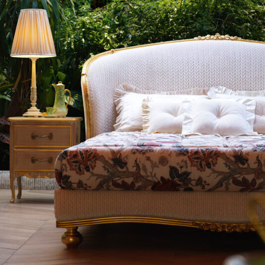 A luxurious bedroom features a Classic Style Antique Gold Winged Bed with an ornate, gold-trimmed headboard and a floral-patterned bedspread adorned with multiple pillows. To the left of the bed is a matching gold-accented nightstand with a lamp and decorative items, set against a lush green backdrop.