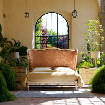 A luxurious bedroom highlights the Classic Style Tall Winged Bed, adorned with a large, ornate headboard featuring a gold damask pattern and draped in a yellow bedspread. Flanking the bed are two matching nightstands, each topped with a lamp. The area is surrounded by lush green plants, and large windows allow abundant natural light to enhance the classic ambiance.