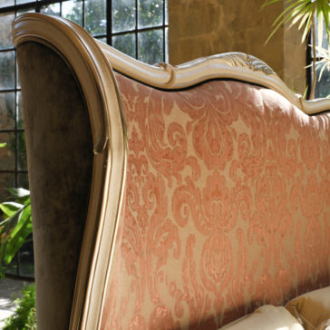 A close-up view of the **Classic Style Tall Winged Bed** showcases its upholstered headboard adorned with intricate red and gold damask patterns, framed by carved wood. Placed near large windows, the bed allows natural light to stream in, highlighting its luxurious texture and design. Green plants are visible in the background.