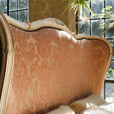 A close-up of the Classic Style Tall Winged Bed features an ornate headboard with a gold frame and intricate pink and gold upholstery. Positioned near a window with black metal frames, sunlight streams in to highlight the detailed design of the headboard.