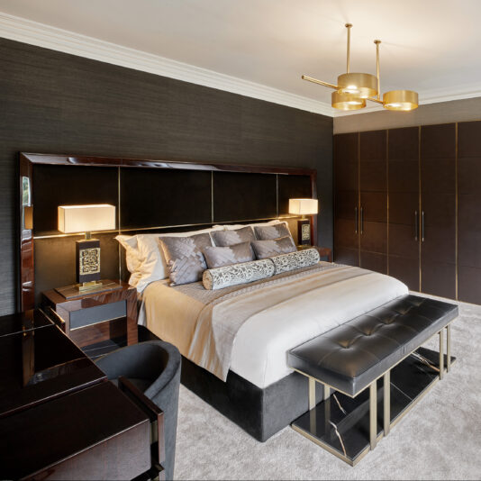A luxurious bedroom featuring a king-sized bed with an ornate headboard, adorned with multiple decorative pillows and cushions. Two matching nightstands with lamps, a cushioned bench at the foot of the bed, sleek dark wardrobes, and a stunning Contemporary Brushed Gold Triple Suspension Light complete the decor.