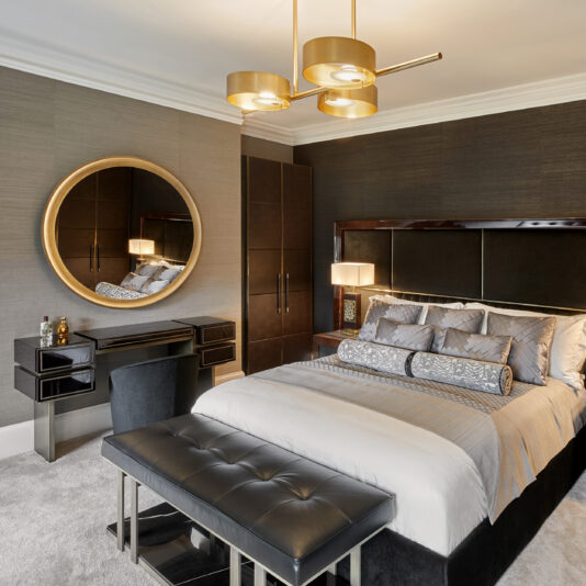 A modern bedroom showcasing a bed with silver and gray bedding, a padded headboard, and a black leather bench at the foot. The room is further enhanced by a vanity table with a chair, a large round mirror, an elegant Contemporary Brushed Gold Triple Suspension Light fixture, and dark-toned walls and furniture.