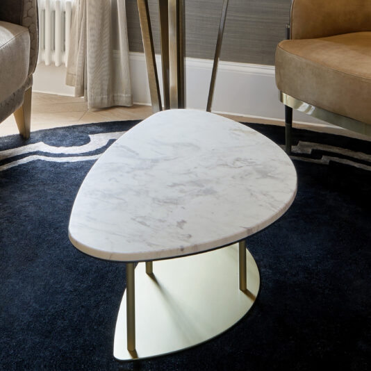 A **Contemporary Marble Coffee Table** with a white, asymmetrical top and a gold metal base stands on a dark blue patterned rug. The surrounding area features beige leather chairs and floor-length curtains partially visible in the background.