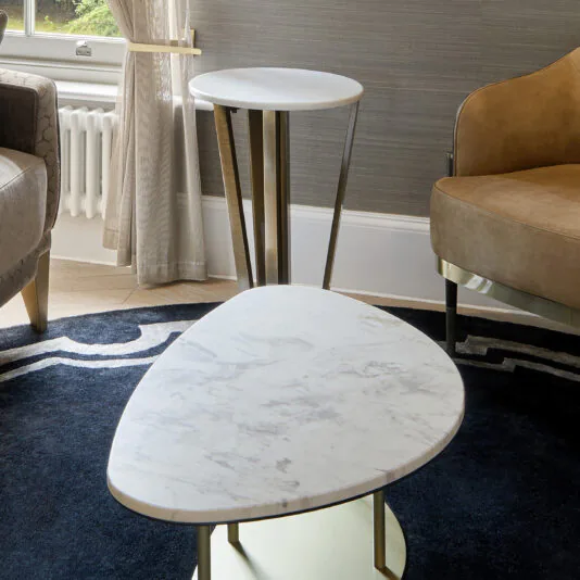 A modern living room with gray walls and a dark blue rug features two beige chairs and a set of Contemporary Marble Coffee Tables, one round and one triangular. A window with cream curtains can be seen in the background.