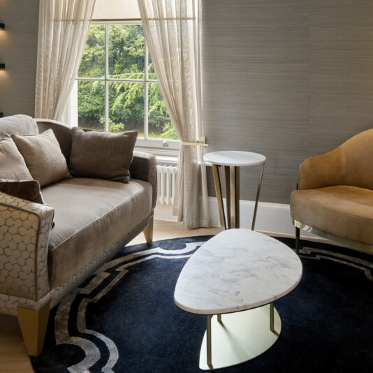 A modern living room features a grey sofa and beige armchair arranged around the Contemporary Marble Coffee Table. A small side table stands by the sofa. The room boasts a large window with sheer curtains, offering a view of lush green trees outside. A blue area rug lies beneath the furniture.