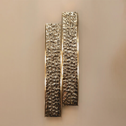 A vertical wall sculpture featuring two textured, metallic rectangular panels of varying lengths, arranged parallel against a neutral backdrop. This exclusive piece mirrors the design of the Exclusive Double Hammered Wall Light with its reflective and intricately hammered finish.