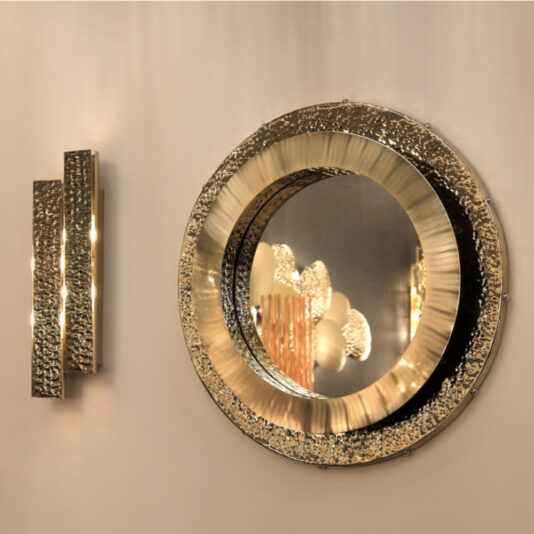 Exclusive Double Hammered Wall Light A refined round mirror with a textured metallic frame hangs on a light-colored wall. Adjacent to it is the Exclusive Double Hammered Wall Light, featuring a similar textured metallic finish and casting a warm glow. The mirror reflects an elegant light fixture with multiple bulbs.