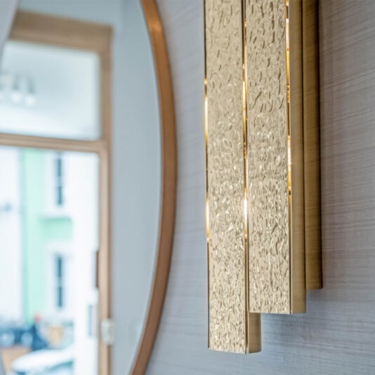 Exclusive Double Hammered Wall Light A close-up of the Exclusive Double Hammered Wall Light mounted beside a large round mirror framed in wood. In the background, a blurred doorway leading outside allows sunlight to gently illuminate the room.