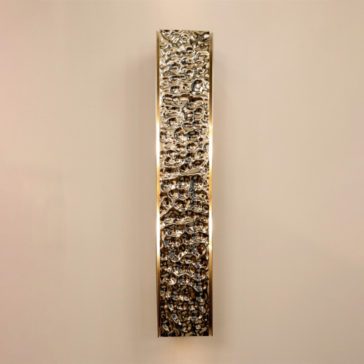 Exclusive Single Hammered Wall Light An Exclusive Single Hammered Wall Light, characterized by its narrow, vertical, rectangular shape and highly textured metallic surface with a polished finish, is mounted on an off-white wall. The gold brass wall sconce appears illuminated from behind, casting a soft glow around its edges.