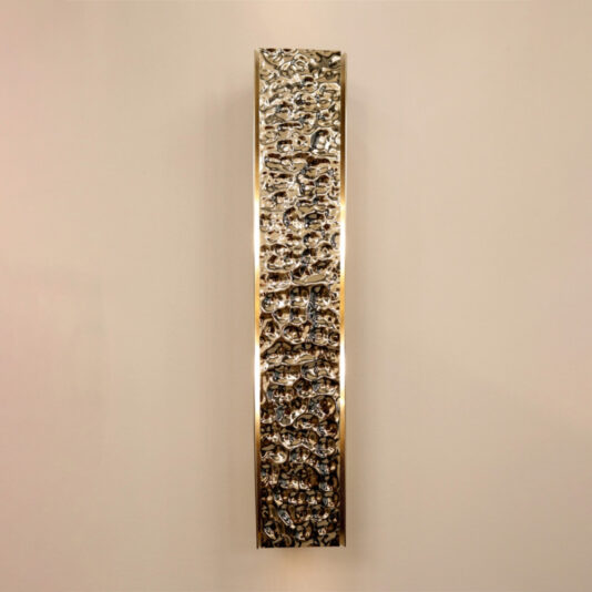 An Exclusive Single Hammered Wall Light, characterized by its narrow, vertical, rectangular shape and highly textured metallic surface with a polished finish, is mounted on an off-white wall. The gold brass wall sconce appears illuminated from behind, casting a soft glow around its edges.