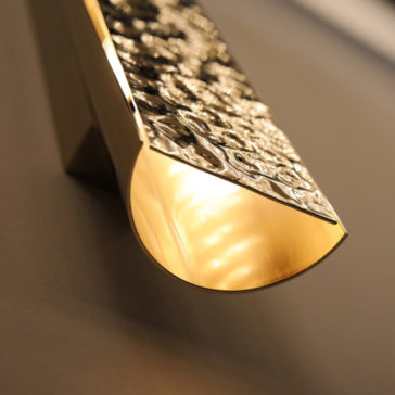 Exclusive Single Hammered Wall Light A close-up of the Exclusive Single Hammered Wall Light with a textured gold brass surface and a curved design. The light emits a warm glow, highlighting the intricate patterns on the fixture. The background wall is slightly out of focus.