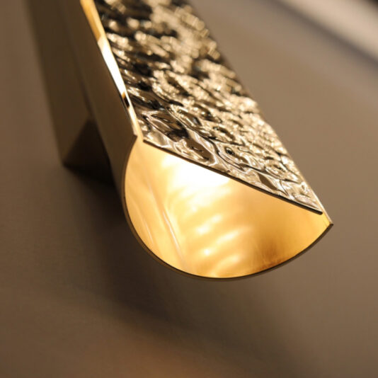 A close-up of the Exclusive Single Hammered Wall Light with a textured gold brass surface and a curved design. The light emits a warm glow, highlighting the intricate patterns on the fixture. The background wall is slightly out of focus.