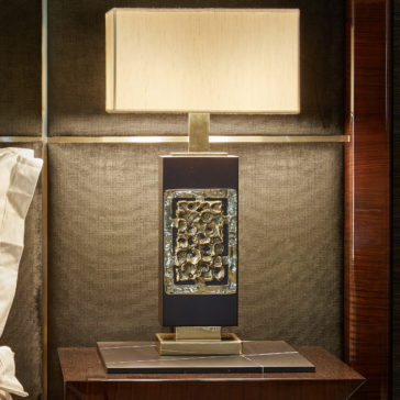 High End Brass Cast Table Lamp The High End Brass Cast Table Lamp, featuring a rectangular base adorned with decorative metallic accents, stands on a dark brown wooden nightstand. Topped with a square beige lampshade, the lamp exudes a modern and artistic look against the textured, dark wall backdrop.