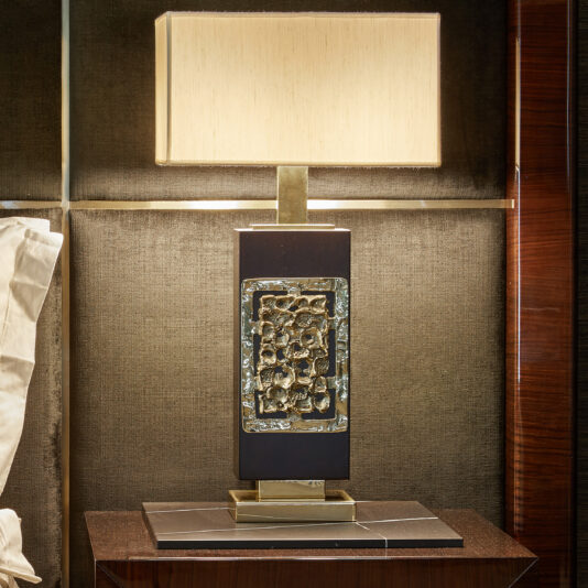 The High End Brass Cast Table Lamp, featuring a rectangular base adorned with decorative metallic accents, stands on a dark brown wooden nightstand. Topped with a square beige lampshade, the lamp exudes a modern and artistic look against the textured, dark wall backdrop.