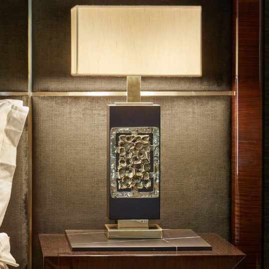 The High End Brass Cast Table Lamp, featuring a rectangular base adorned with decorative metallic accents, stands on a dark brown wooden nightstand. Topped with a square beige lampshade, the lamp exudes a modern and artistic look against the textured, dark wall backdrop.