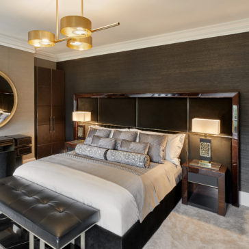 High End Brass Cast Table Lamp A modern bedroom features a large bed with gray and silver bedding, flanked by two nightstands each adorned with a High End Brass Cast Table Lamp. A tufted bench is positioned at the foot of the bed. The room is decorated with dark textured walls, a round mirror, and elegant gold pendant lighting.
