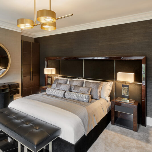 A modern bedroom features a large bed with gray and silver bedding, flanked by two nightstands each adorned with a High End Brass Cast Table Lamp. A tufted bench is positioned at the foot of the bed. The room is decorated with dark textured walls, a round mirror, and elegant gold pendant lighting.
