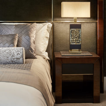 High End Brass Cast Table Lamp A cozy bedroom with a neatly made bed featuring textured gray pillows and a light beige comforter. Next to the bed is a sleek wooden nightstand adorned with the High End Brass Cast Table Lamp and a decorative stone object. The lamp emits a warm glow, creating a tranquil ambiance.