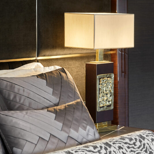 In a close-up shot, a bed with a tufted headboard is adorned with grey patterned pillows and an animal print blanket, while on the nightstand beside it sits the High End Brass Cast Table Lamp, featuring a rectangular shade and an ornate base. The room exudes luxurious elegance.