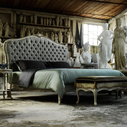 This luxurious bedroom features the High End Classic Style Button Upholstered Bed with its ornate headboard and footboard, set in a rustic room adorned with classical sculptures. The space boasts a large window, weathered floors, and shelves displaying numerous statues, all contributing to an artistic and elegant ambiance that exudes high-end classic style.