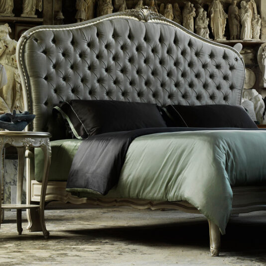 An elegant bedroom features the High End Classic Style Button Upholstered Bed, showcasing its dark tufted buttons. The bed is adorned with green bedding and complemented by black pillows. Beside the bed sits a classic nightstand, enhancing the room's sophisticated ambiance. Statues decorate the background.