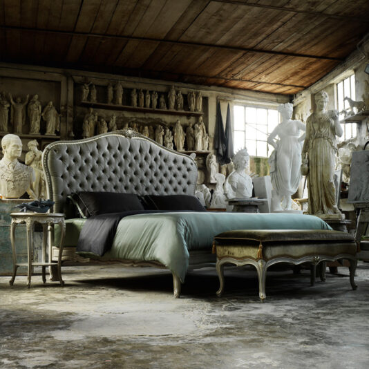A rustic bedroom showcases a High End Classic Style Button Upholstered Bed, featuring a large tufted headboard, black pillows, and a green duvet. At the foot of the bed sits an upholstered bench. The room is adorned with numerous classical sculptures and statues set against weathered walls and beneath a wooden ceiling.