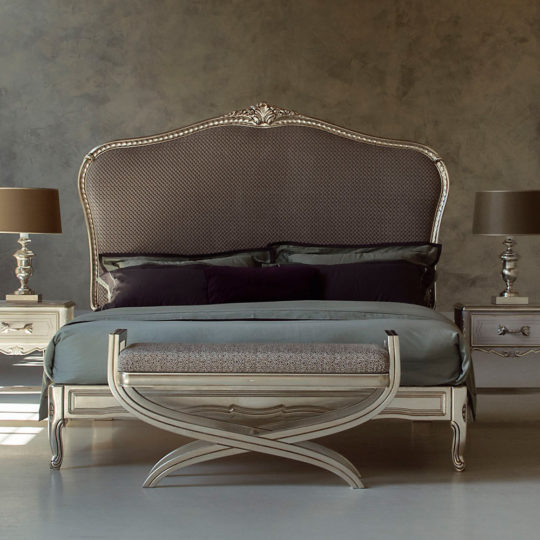 A high-end bedroom features the luxurious High End Classic Style Upholstered Bed with a grand headboard adorned with ornate detailing, epitomizing classic style. The bed is neatly made with muted gray and green bedding. A cushioned bench sits at the foot of this elegant upholstered bed, while matching nightstands with lamps flank each side.