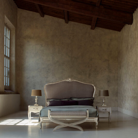 A stylish bedroom showcasing the High End Classic Style Upholstered Bed, which includes a large, ornate headboard and decorative silver frame. Two nightstands with matching table lamps flank the bed, while daylight streams through a large window to the left, softly illuminating the room. The slanted wooden ceiling adds rustic charm to the setting.